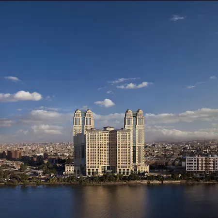فندق فيرمونت نايل سيتي القاهرة المظهر الخارجي الصورة The photo shows a large, modern building complex that appears to be situated by a river, likely in an urban environment. The structure features tall towers, with a distinctive architectural design, and is surrounded by a cityscape that includes other